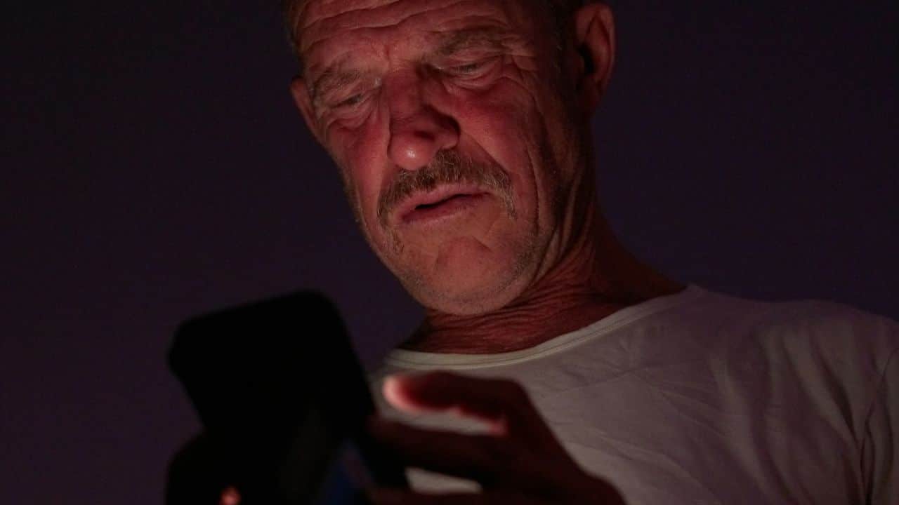 A worried man looking down at his phone in a dark room.