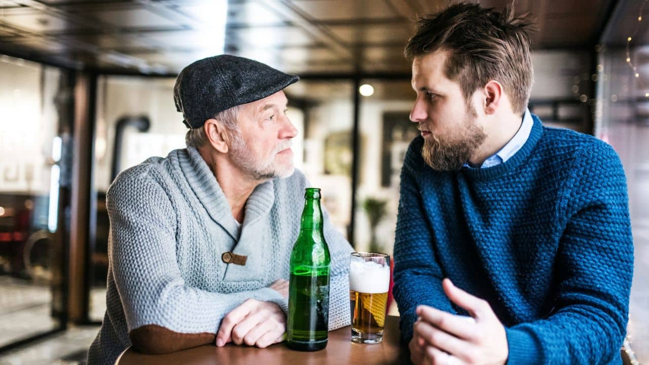 An older man and a younger man drinking beer and talking in a bar.