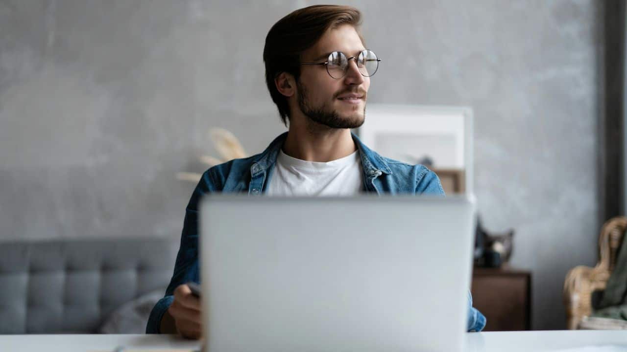 A handsome man in a denim jacket is smiling while looking away from his laptop.