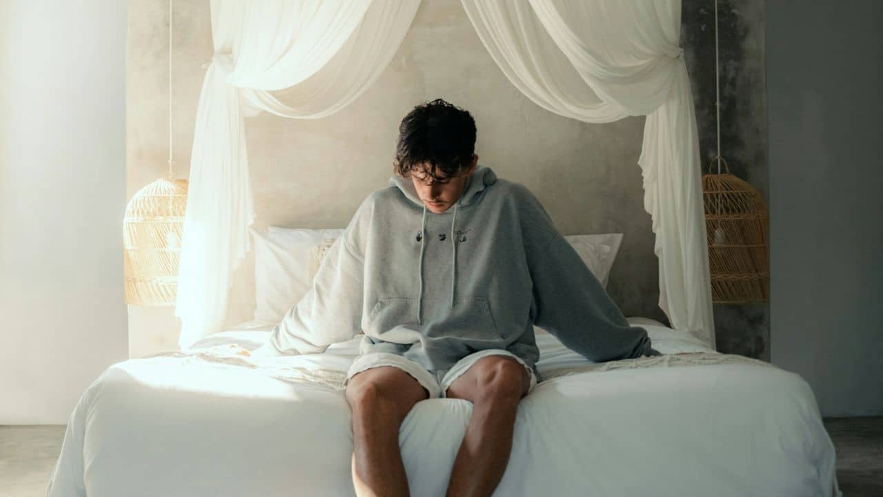 A depressed man is sitting alone on a bed with his head bowed.