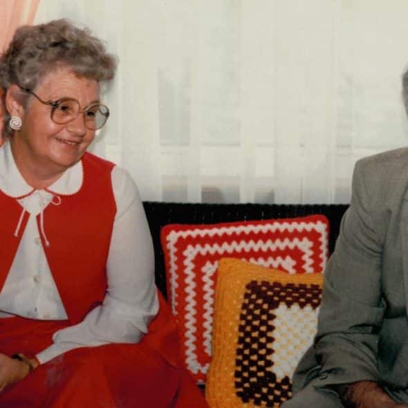 An elderly couple is sitting on a couch. The woman is smiling and holding a teacup, while the man is looking to the side.