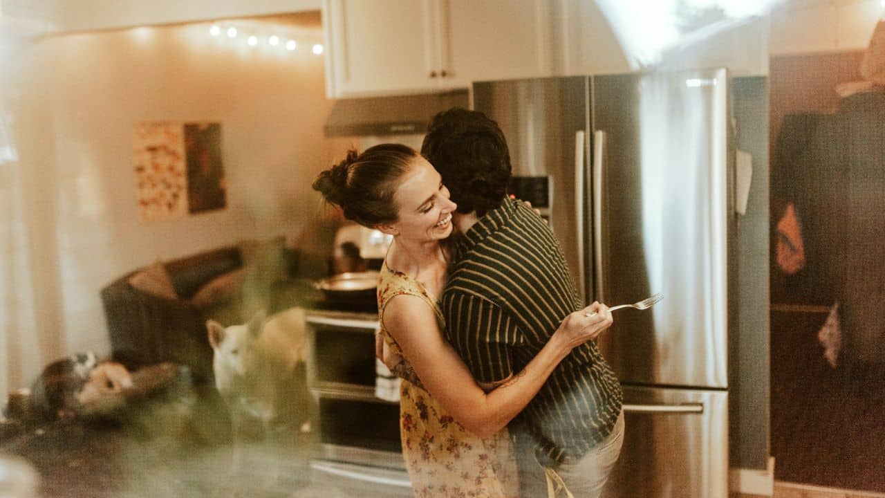 A smiling couple is hugging in a kitchen with a dog.