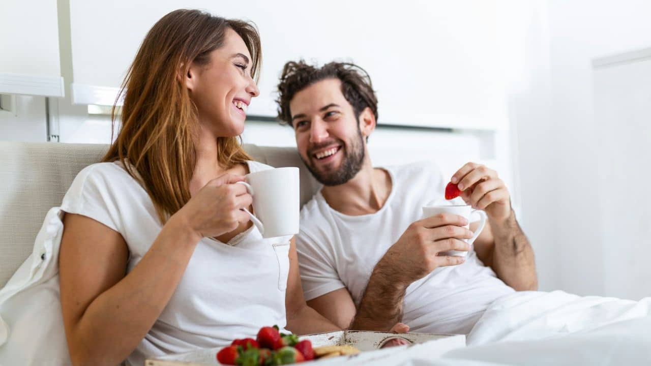 A happy couple smiling at each other while having coffee and strawberries in bed.