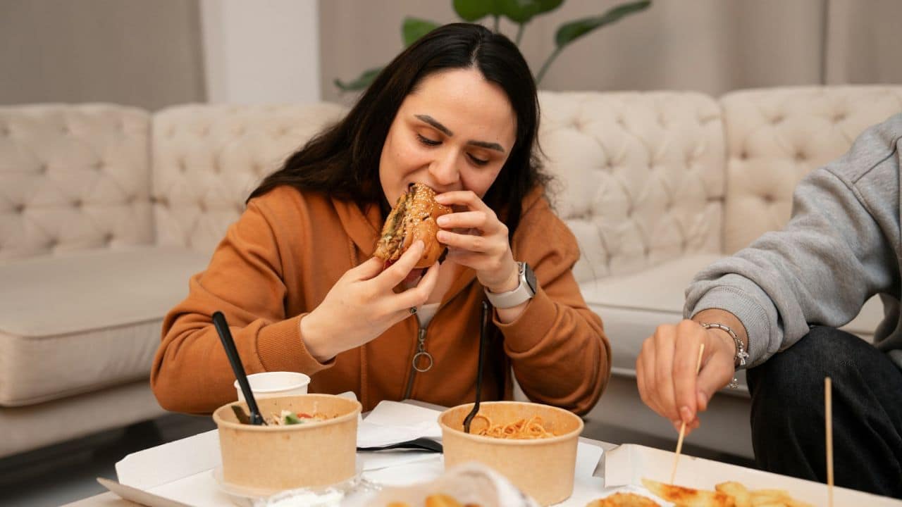A woman in an orange hoodie is eating a large sandwich.