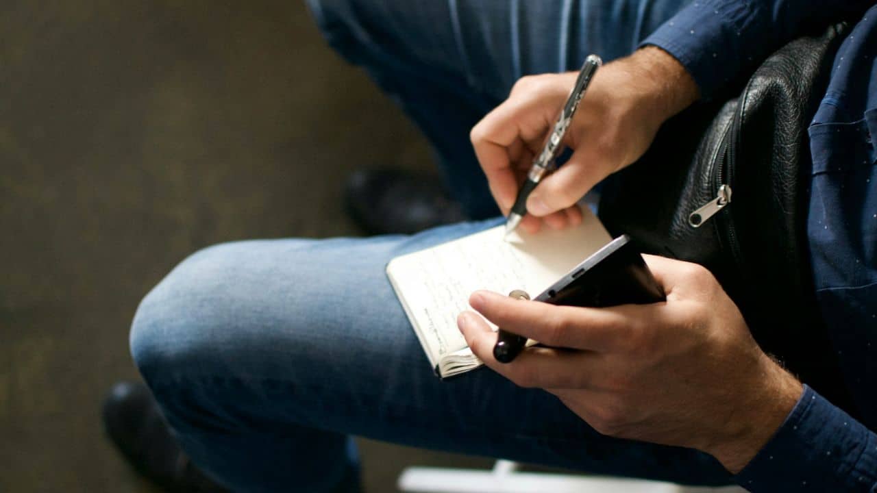 A person is sitting down and writing on a small notepad with a pen.