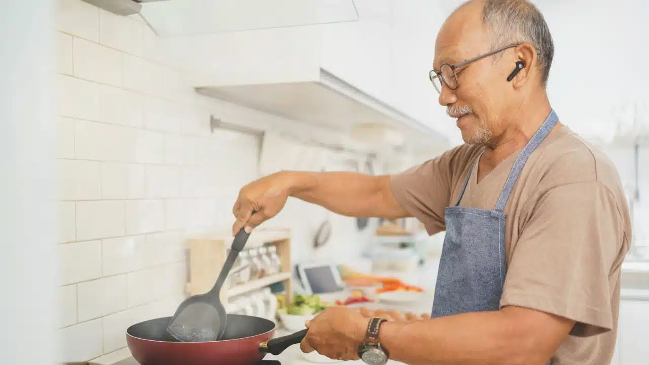 A happy senior man wearing glasses and an apron cooking in a kitchen.