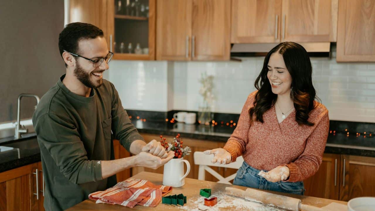 A happy couple smiling at each other while baking in a kitchen.