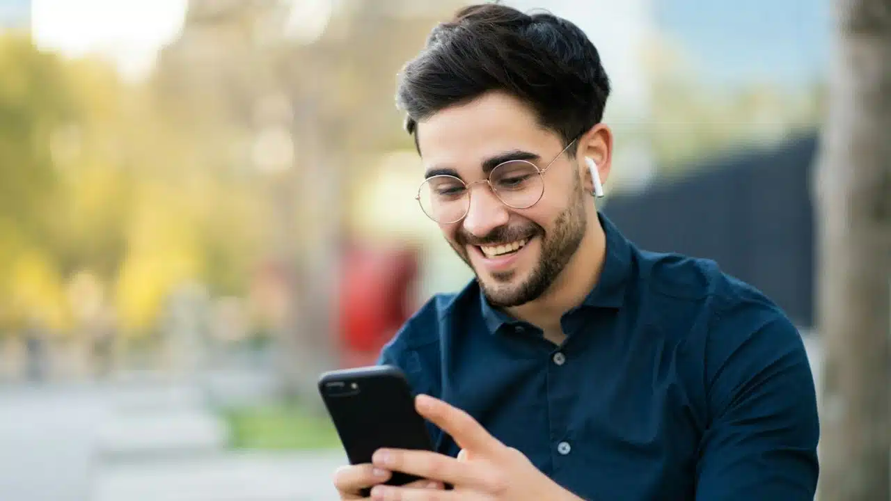 A happy man wearing glasses and earbuds, looking at his phone.
