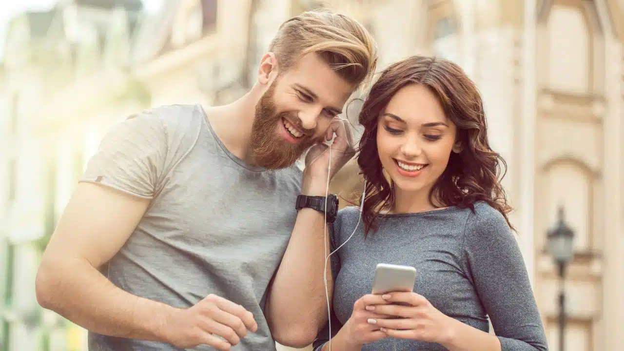 A couple is sharing earphones, smiling, and looking at a smartphone.