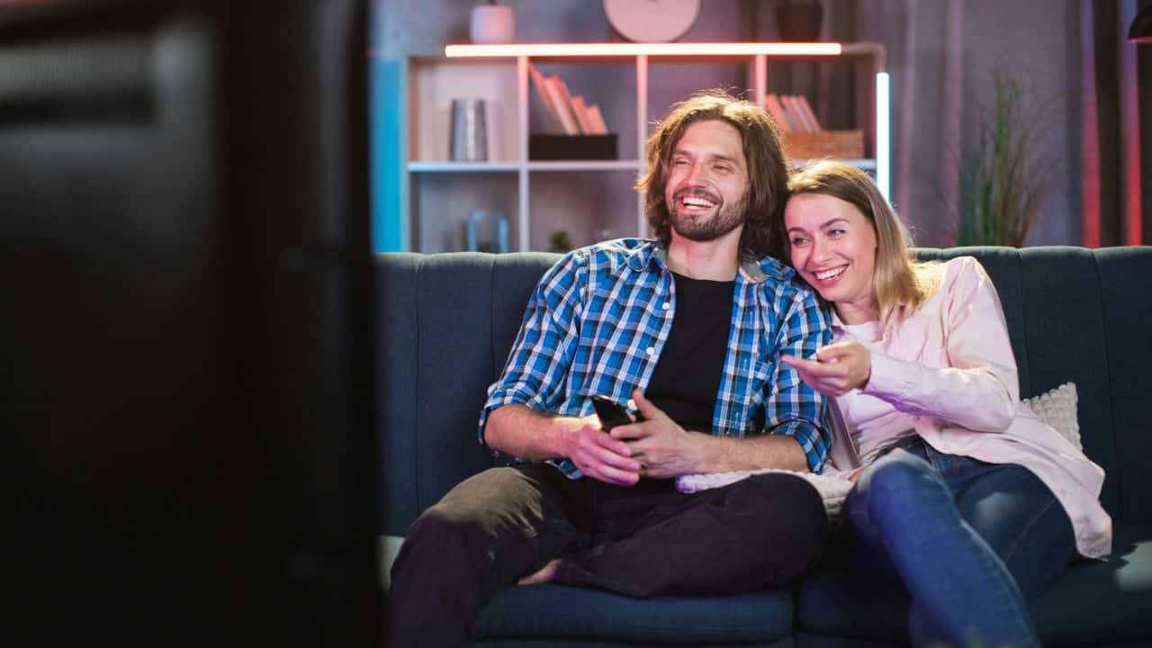 A smiling couple sitting on a couch, watching a show and pointing at the TV.