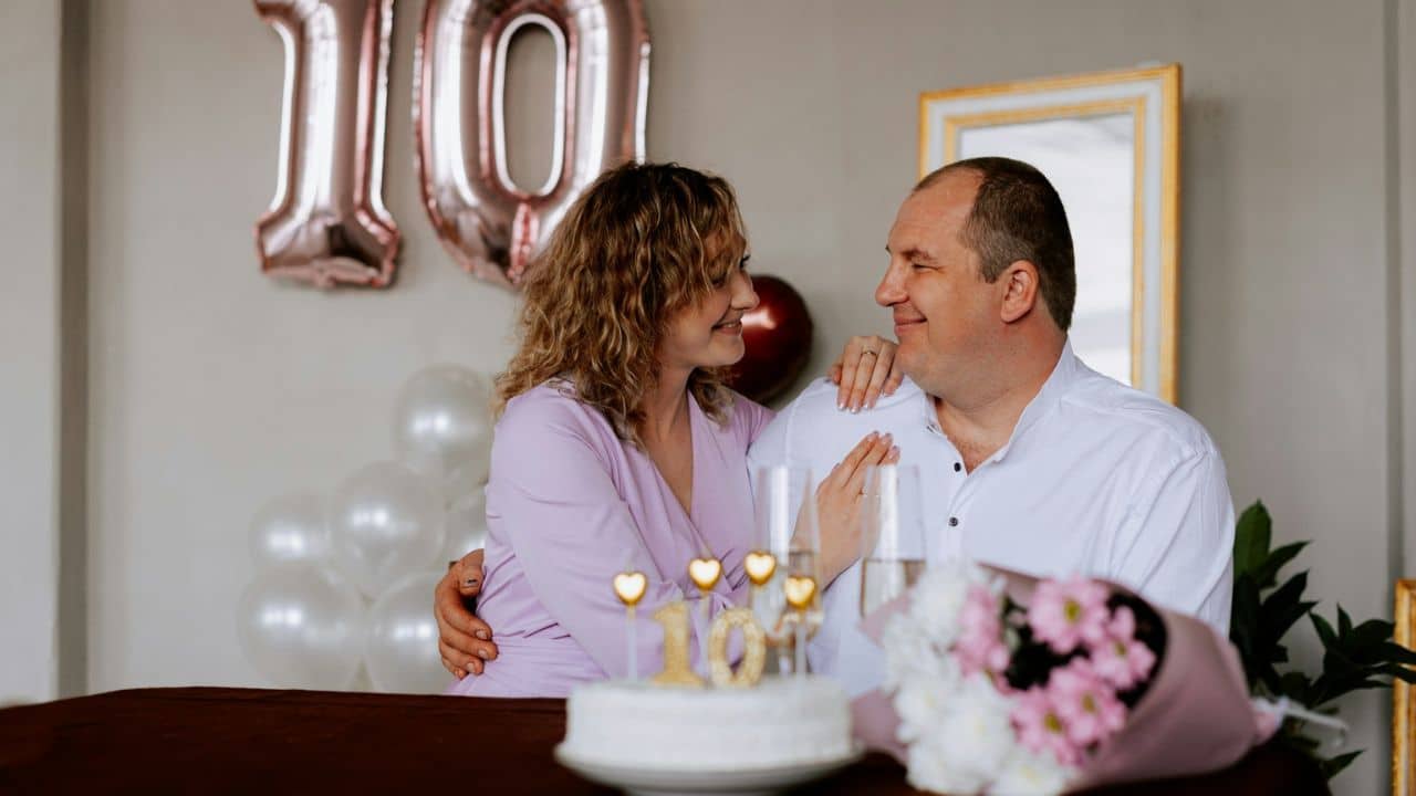 A smiling couple celebrating with a cake, glasses, balloons, and flowers.