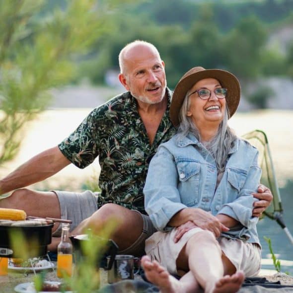 A smiling senior couple sitting by a lake, hugging while having a picnic.