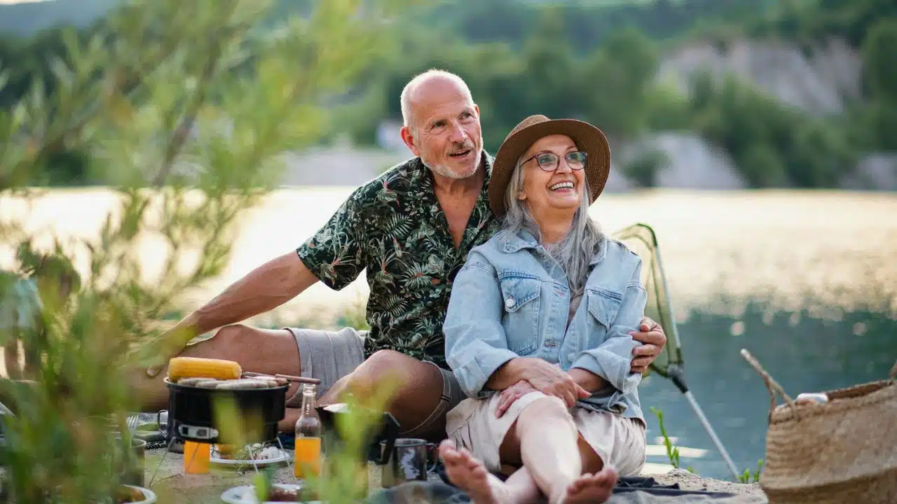 A smiling senior couple sitting by a lake, hugging while having a picnic.