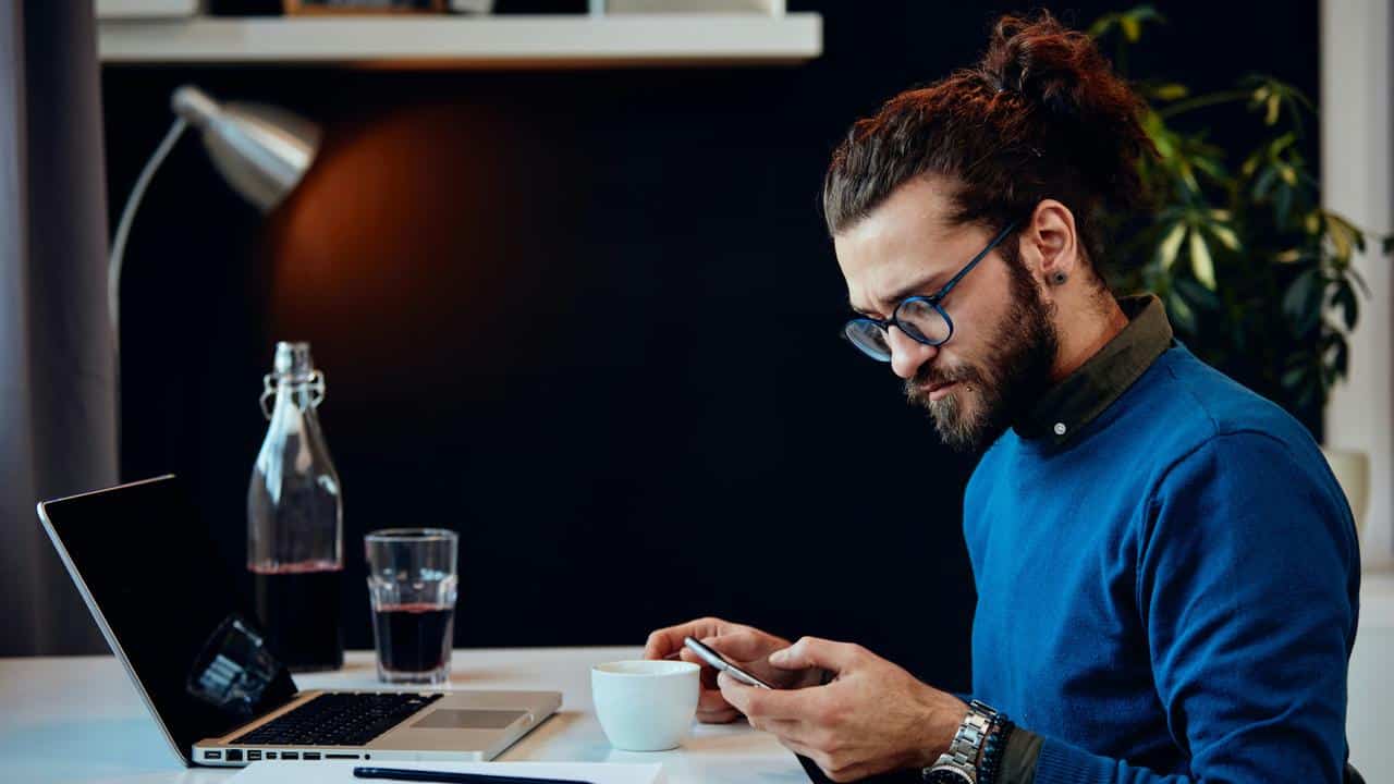 A man using his smartphone at a desk with a laptop and coffee..