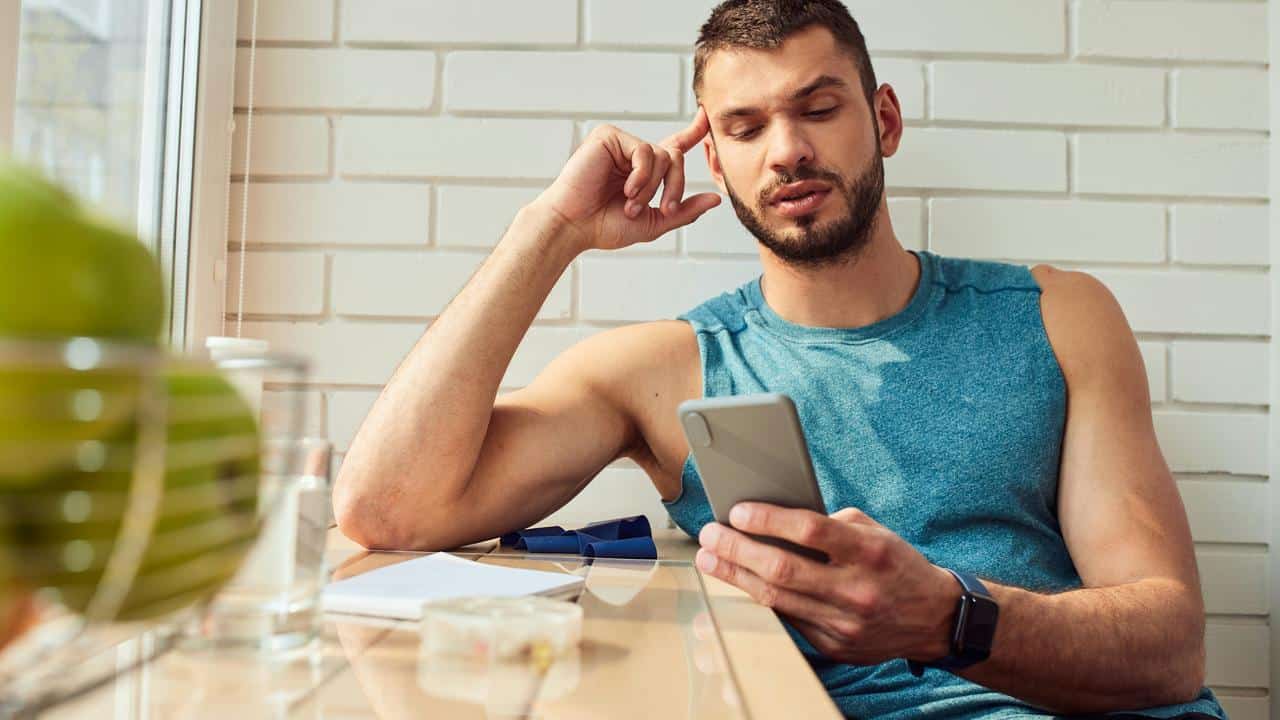 A man in a sleeveless shirt looking at his smartphone by a window.