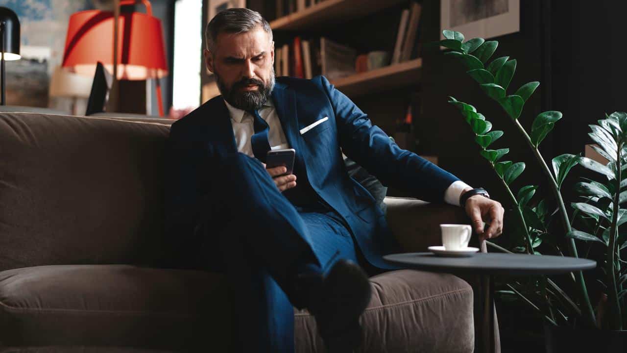 A man in a suit sitting on a couch looking at his smartphone.