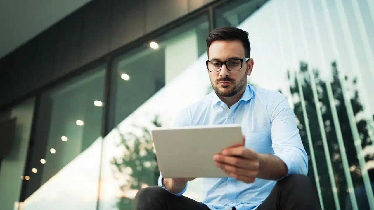 A man in glasses using a tablet outside a building.