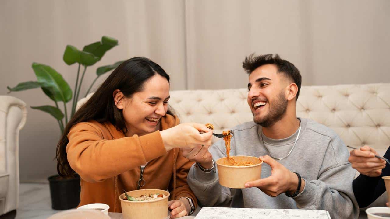 A smiling couple sharing noodles while eating together indoors.