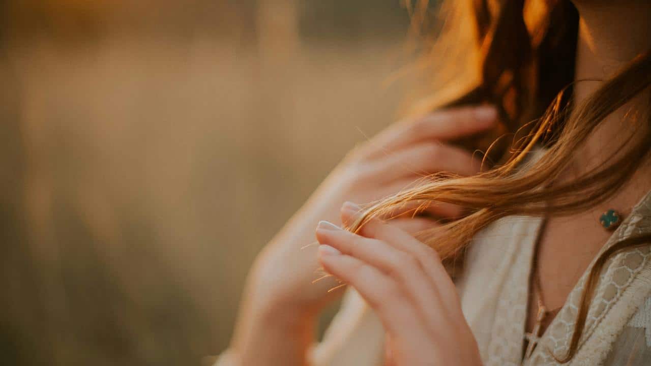 A close-up of a woman’s hands gently touching her hair in warm sunlight.