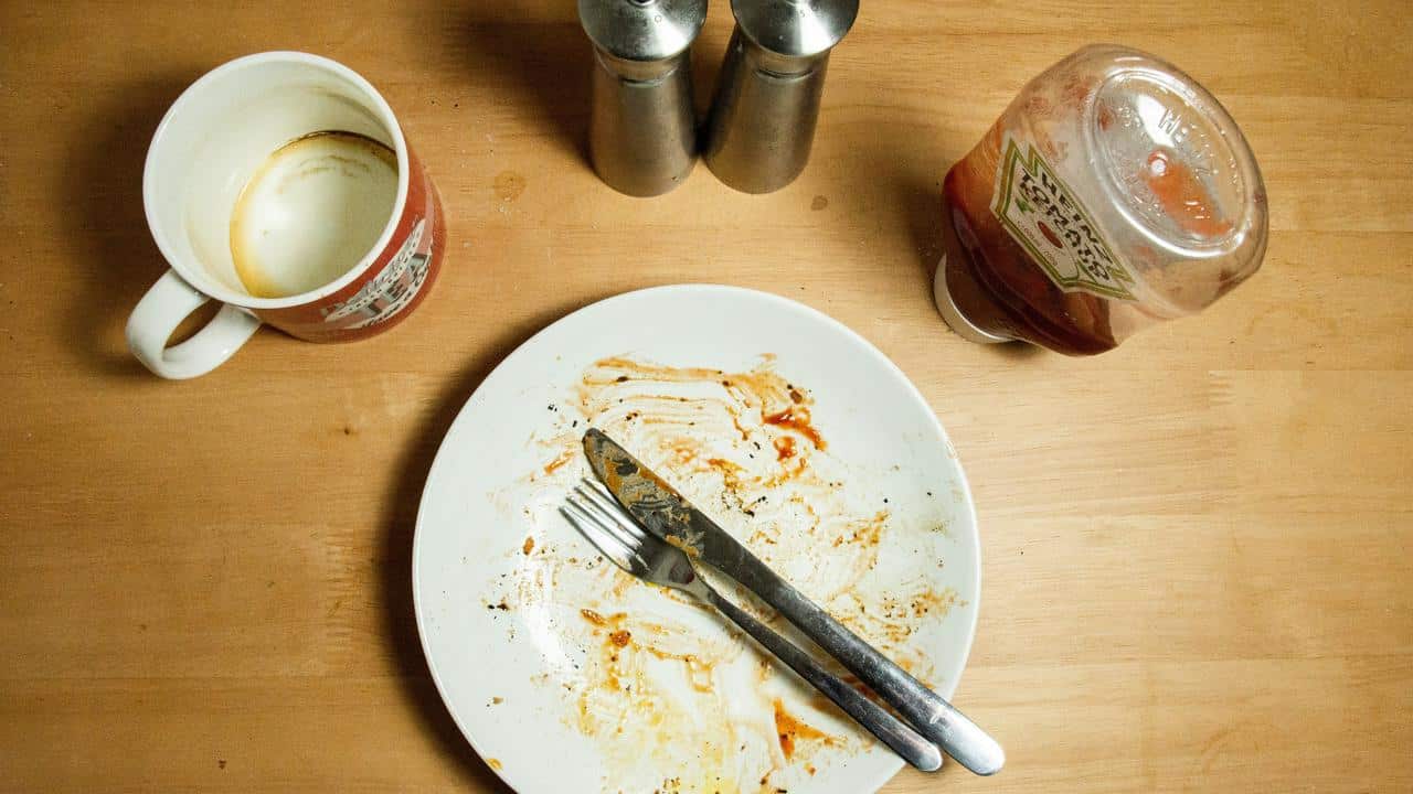A dirty plate with cutlery, an empty mug, and a ketchup bottle on a table.