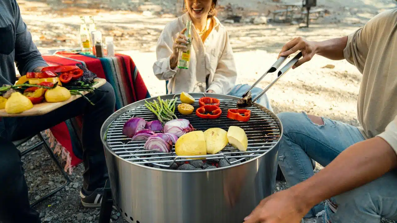 A group of friends grilling vegetables outdoors on a round barbecue grill.