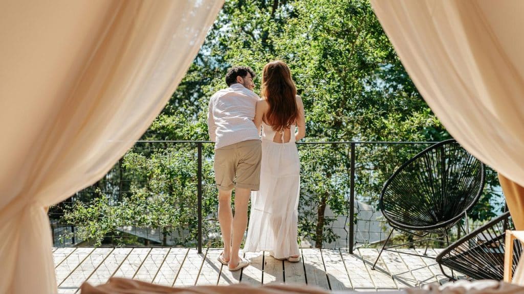 A couple stands on a balcony, framed by curtains, enjoying a peaceful view of the trees.