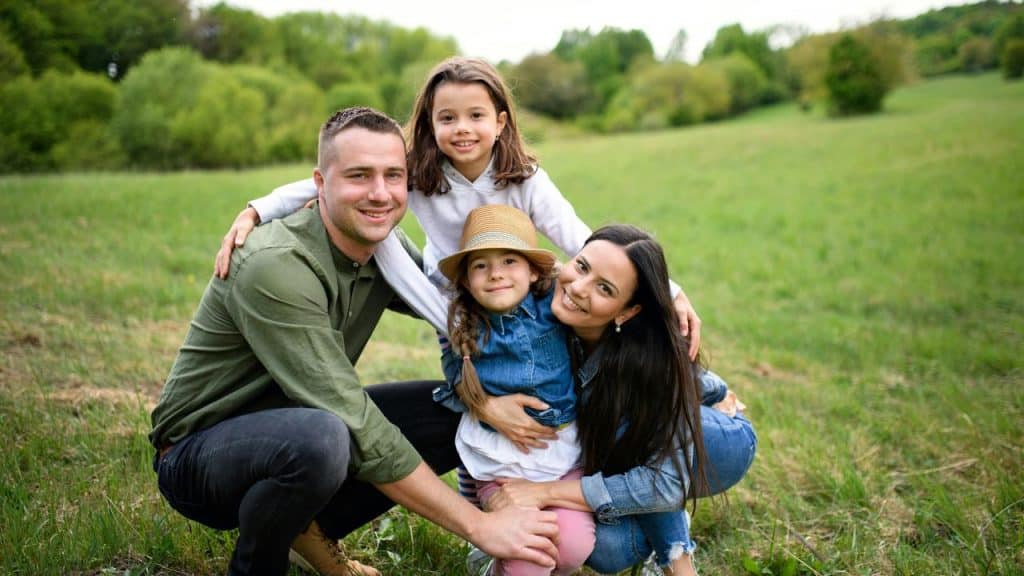 A smiling family with two children poses together in a grassy field.