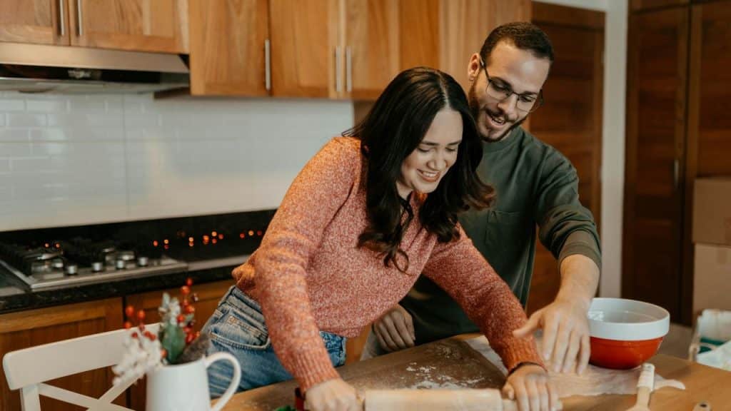 A couple smiles while rolling dough together in the kitchen.