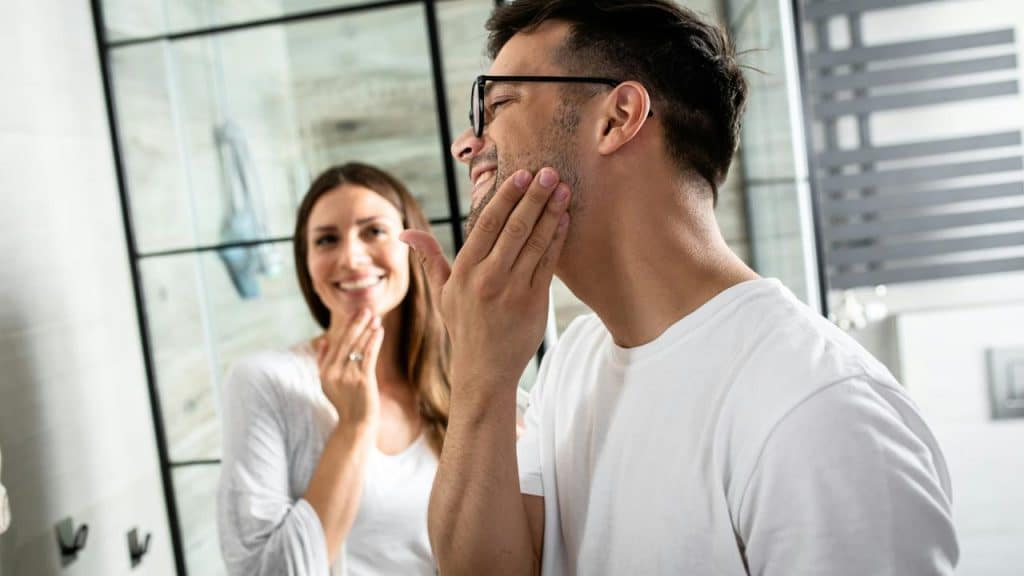 A couple smiles in the bathroom while applying skincare.