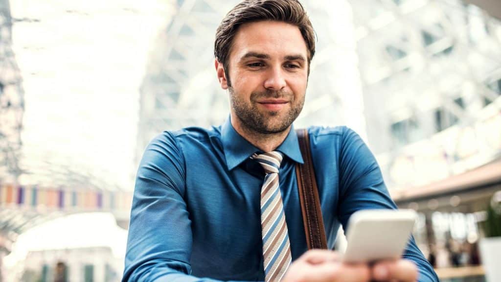 A man in a blue shirt and tie looks at his phone with a slight smile.