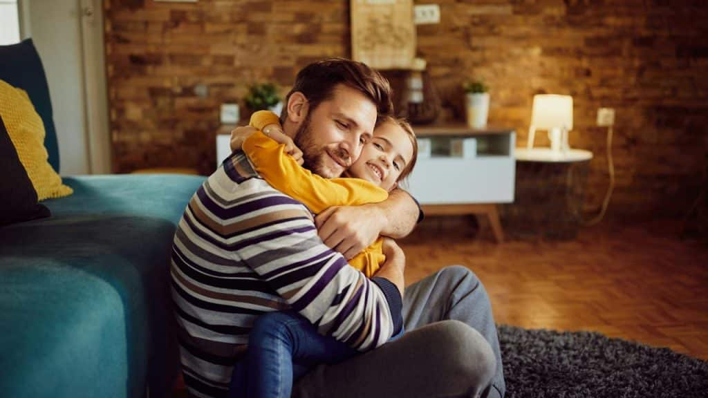 A father hugs his smiling daughter while sitting on the floor.