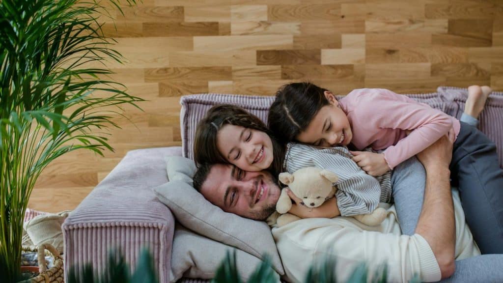 A father cuddles on the couch with his two smiling daughters.