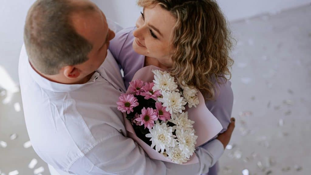 A couple embraces while holding a bouquet of pink and white flowers.