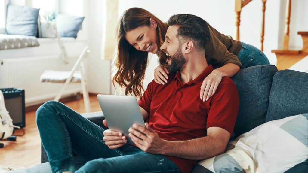 A couple laughs together while looking at a tablet on the couch.