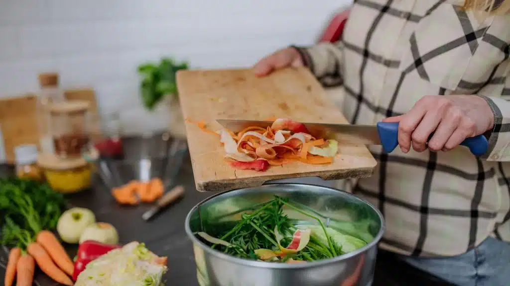 A person scraping vegetable peels from a cutting board into a metal bowl.