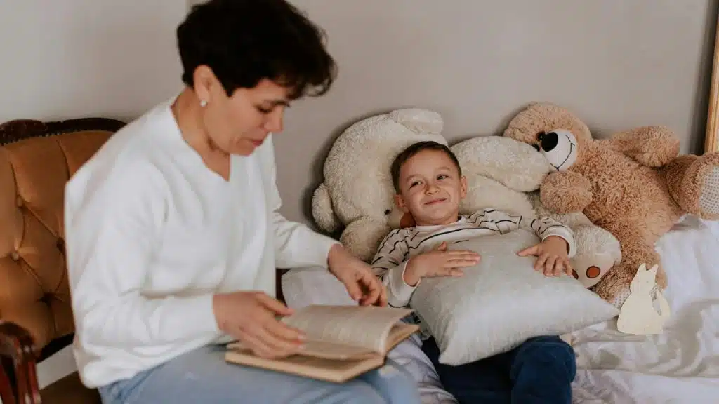 A woman reading a book to a child lying in bed with stuffed animals.