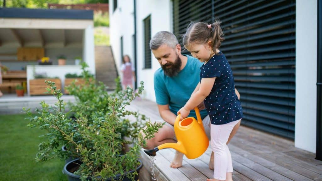 A father helping his daughter water plants with a watering can.