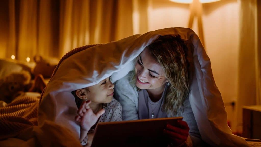 A mother and daughter smiling under a blanket while looking at a tablet.