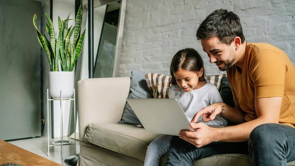 A father and daughter sitting on a couch smiling while looking at a laptop.