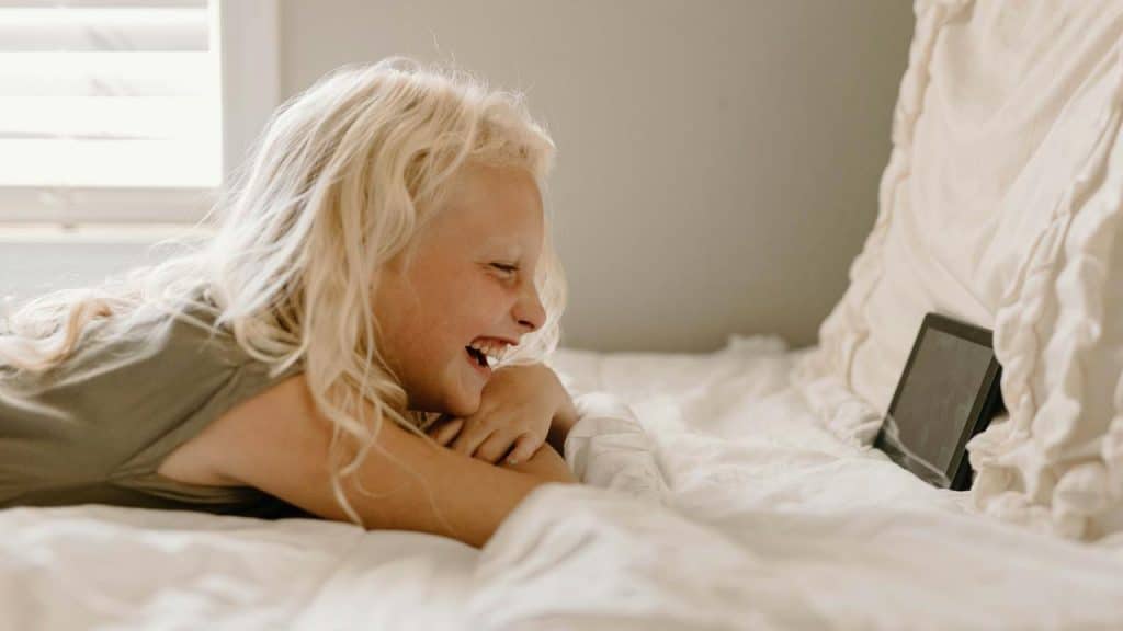 A smiling child lying on a bed while watching a tablet.