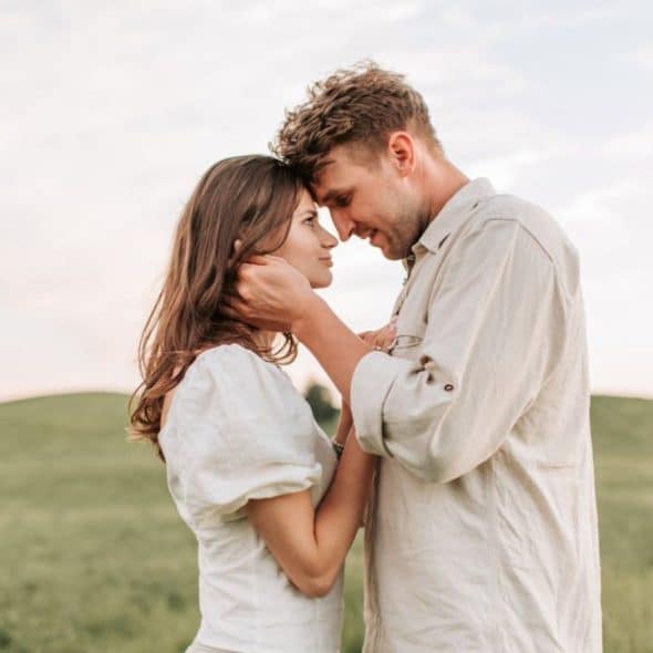 A couple stands in a field, touching foreheads and looking into each other's eyes.