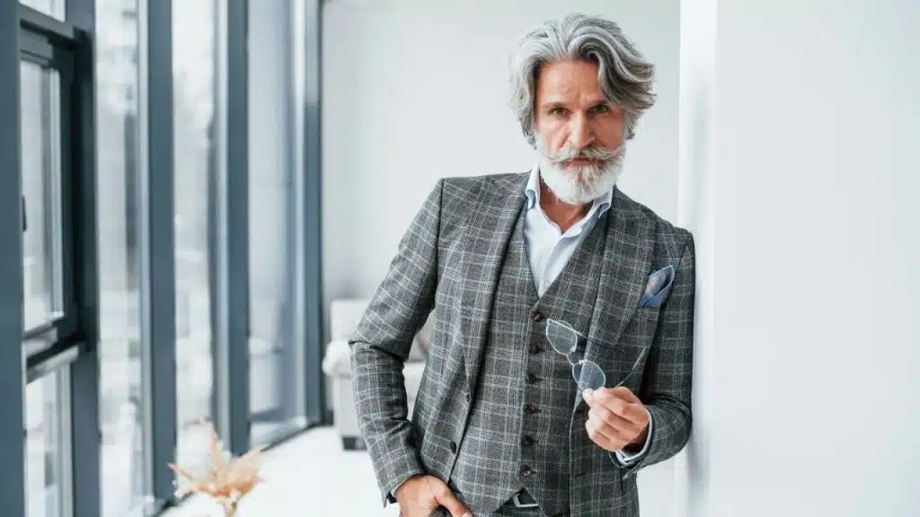 A senior man in a gray plaid suit jacket holding his glasses.