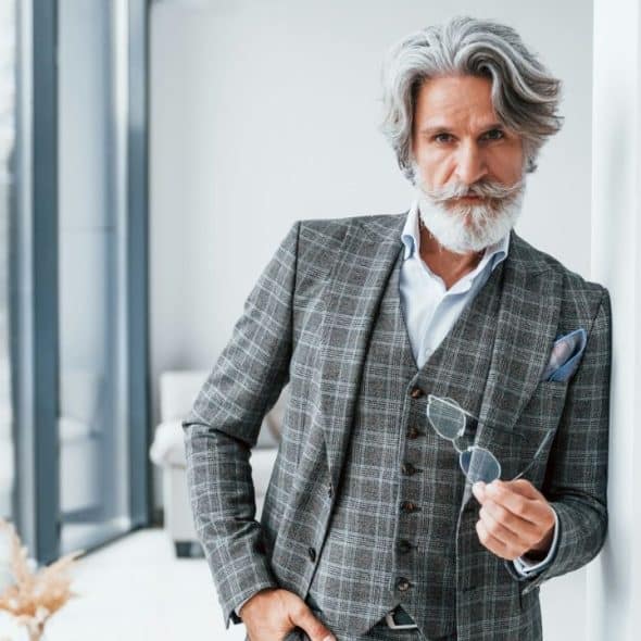 A senior man in a gray plaid suit jacket holding his glasses.