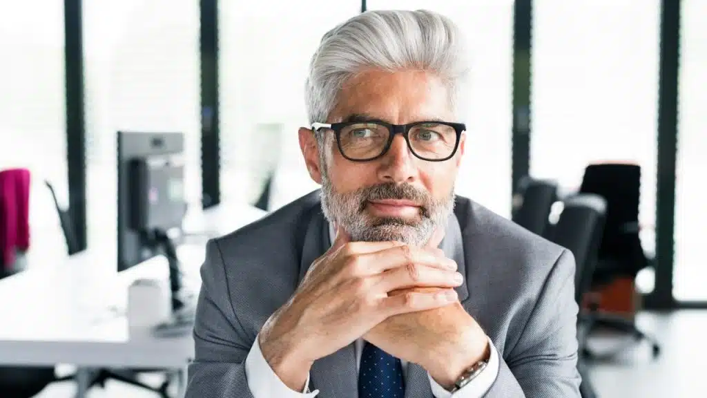 A middle-aged man with gray hair and a beard, wearing glasses and a suit.