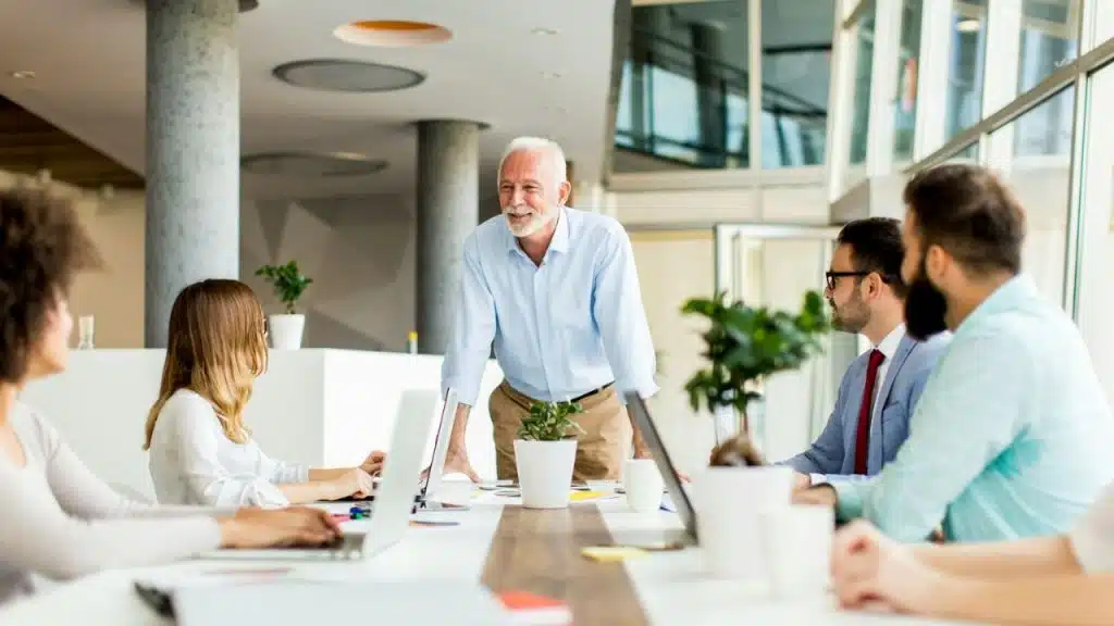 A happy senior man is standing at the head of a table, looking at his colleagues.