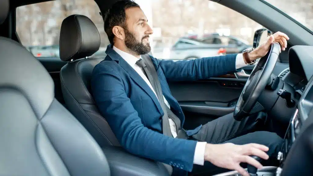 A smiling, well-dressed businessman with a beard sits in the driver's seat of a car.