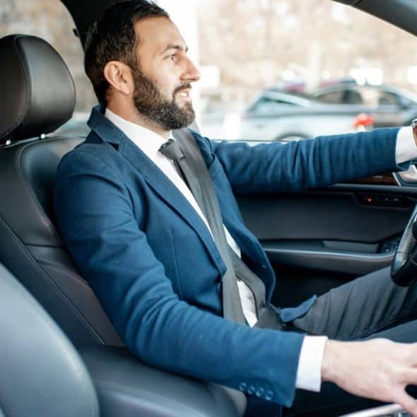 A smiling, well-dressed businessman with a beard sits in the driver's seat of a car.