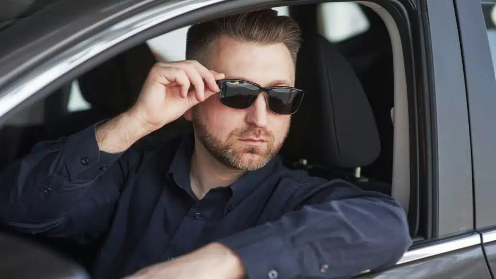 A bearded man in a car, tipping his sunglasses down to look at someone.