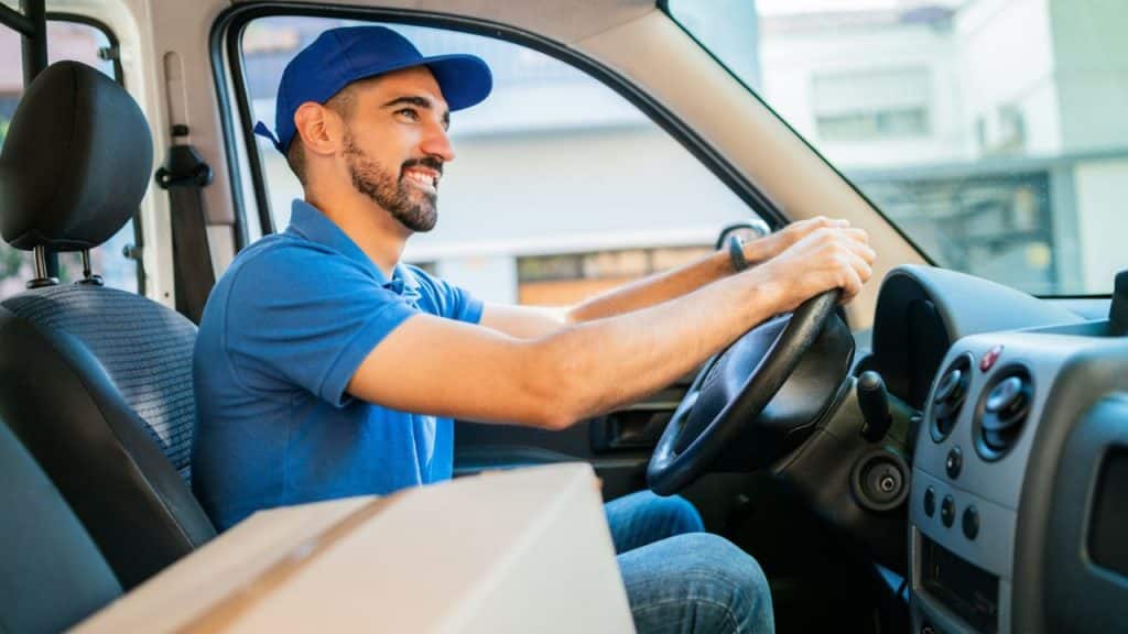 A smiling delivery man in a blue uniform and cap driving a van.