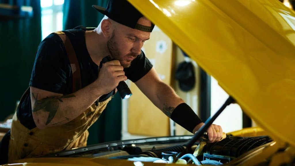 A bald mechanic with tattoos and a cap looks at a car engine with a flashlight.